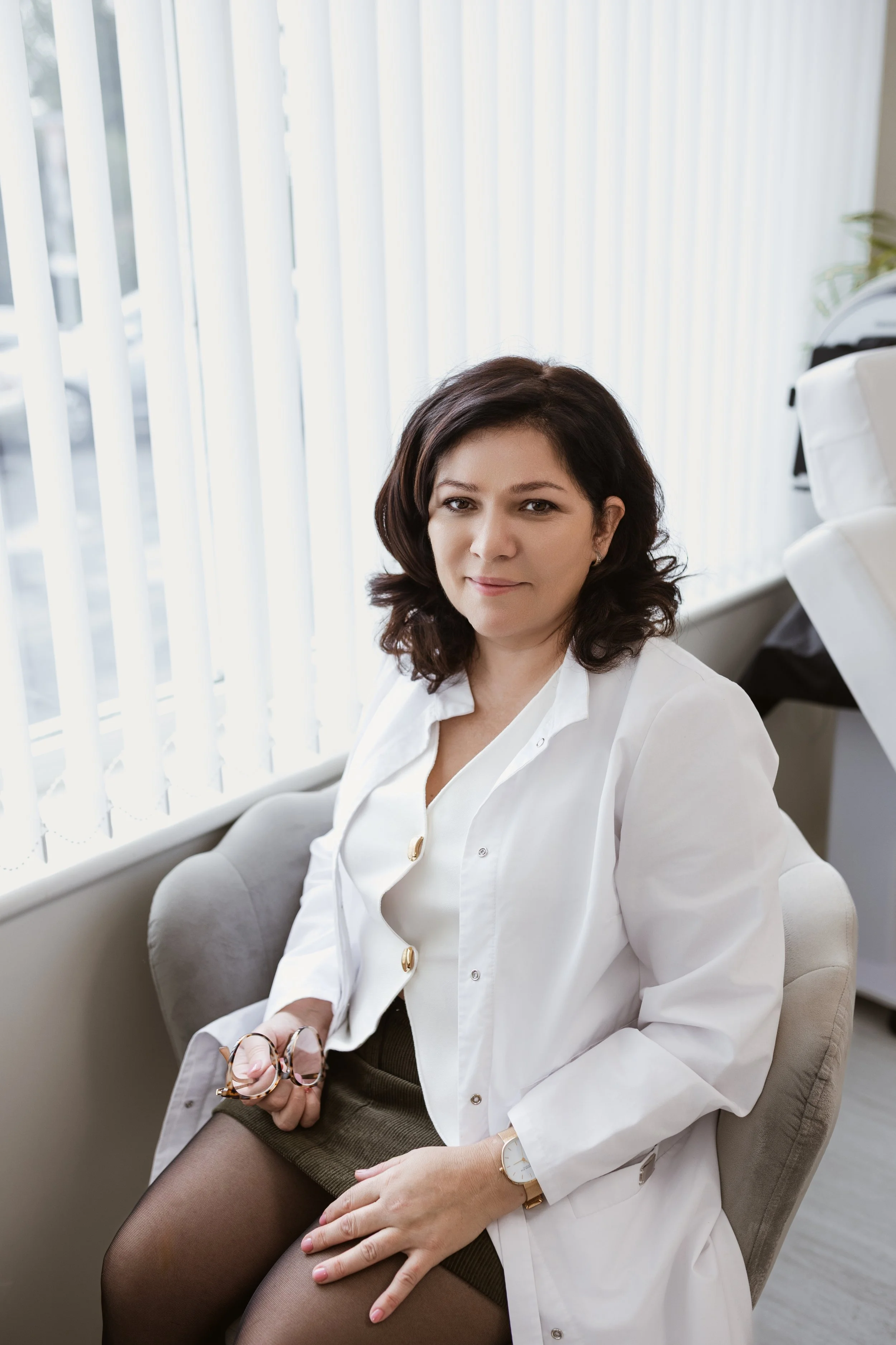 A woman with dark curly hair, wearing a white shirt and a medical white coat, sitting on a beige chair by a window with vertical blinds.