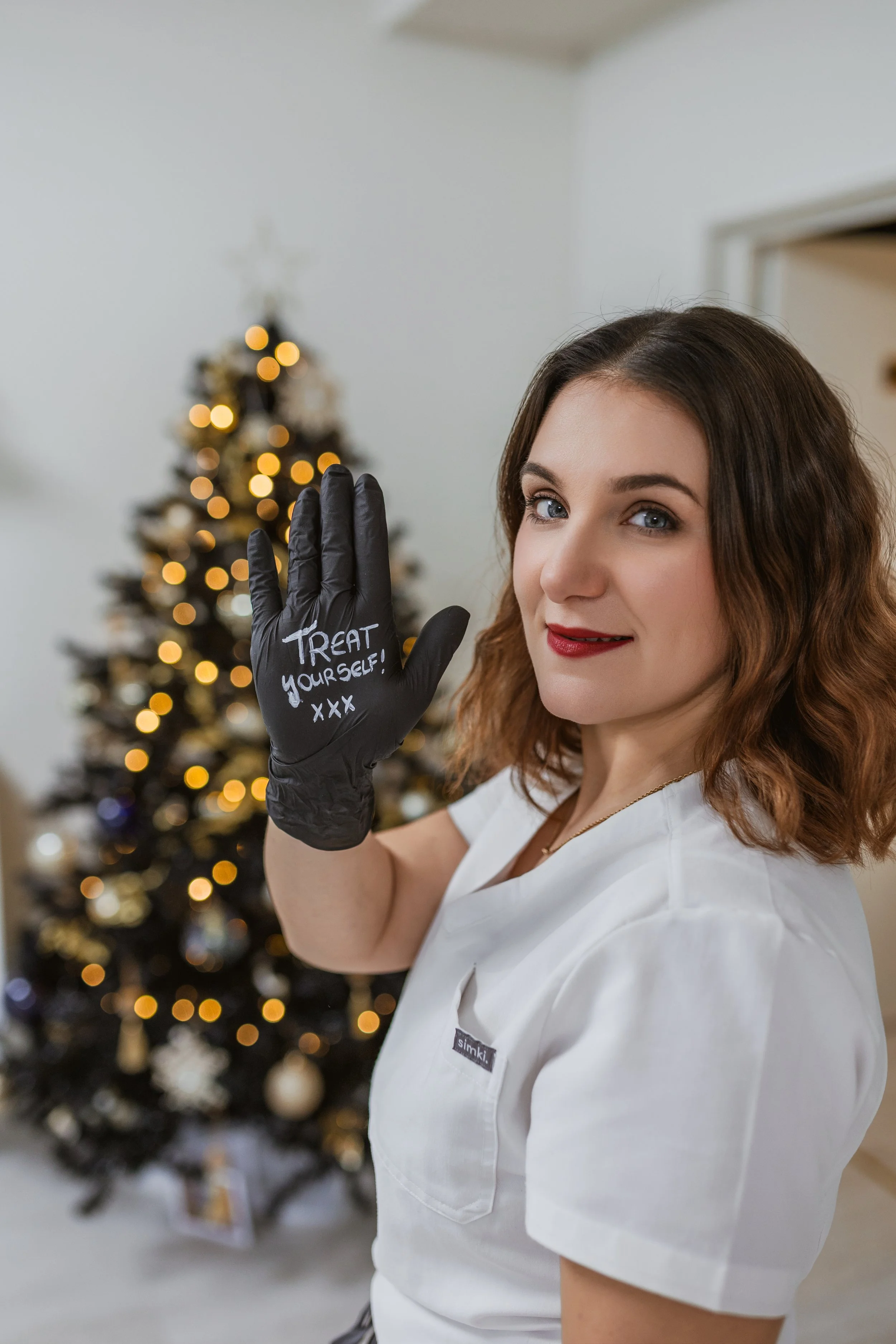 A woman in a white uniform showing a black glove with the message 'TREAT yourself! XXX' written on it, standing in front of a decorated Christmas tree.