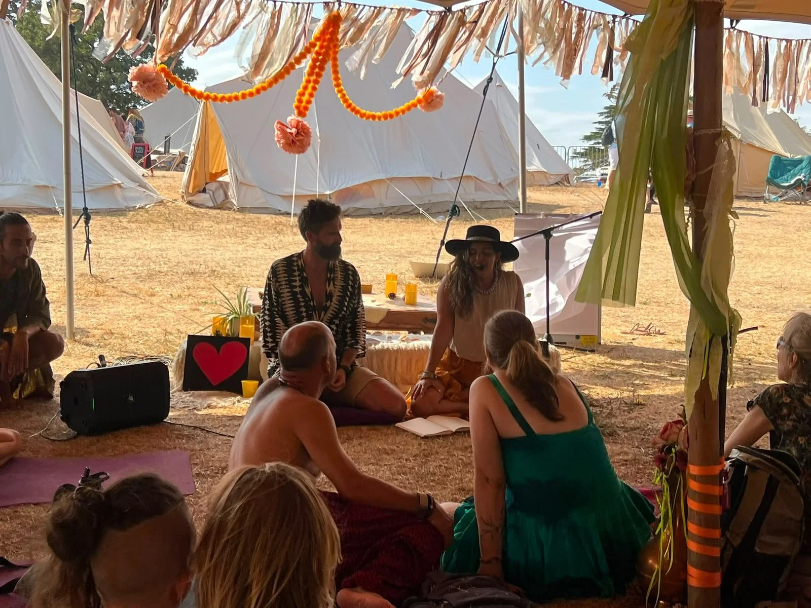 People gathered under a decorated tent, sitting on the ground, engaged in a discussion or performance at an outdoor event, with tents and trees in the background.