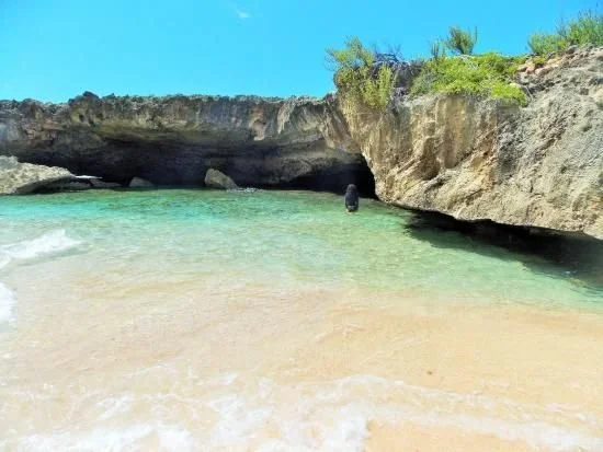 A person standing near a small opening in a rocky coastal formation along a sandy beach, with clear turquoise water and blue sky.