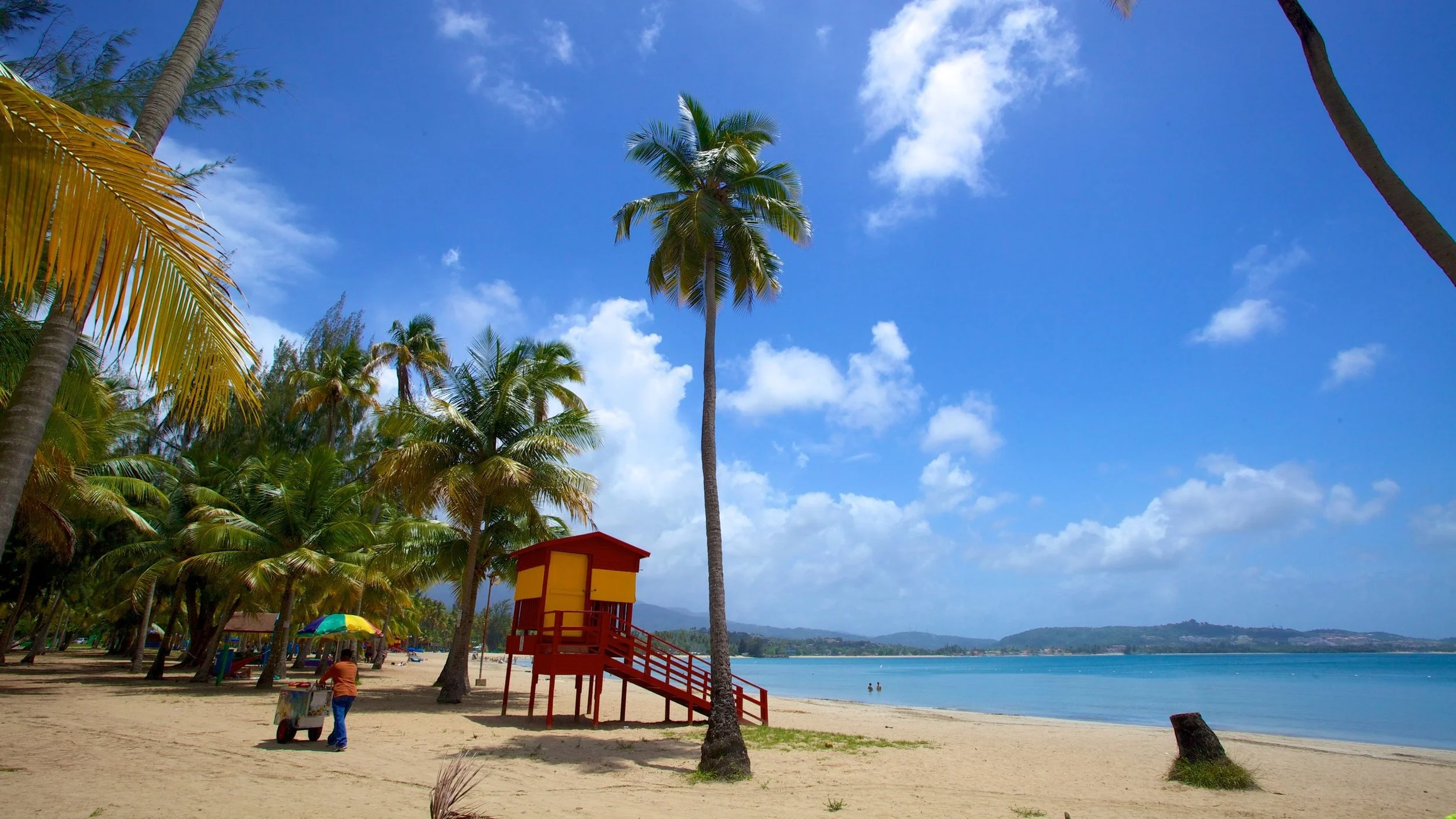 Beach with sandy shore, palm trees, a lifeguard tower painted yellow and red, a vendor pushing cart with an umbrella, and calm ocean waters under a blue sky with scattered clouds.