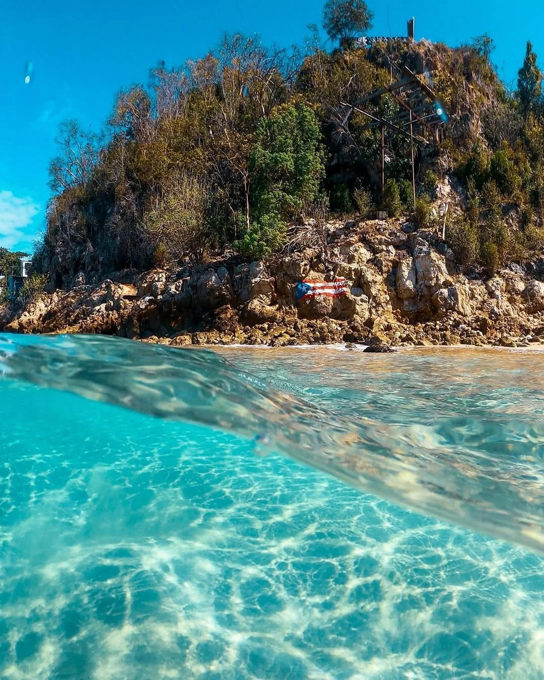 A photo taken partly underwater showing clear blue water in the foreground and rocky shoreline with green bushes and trees in the background. The shoreline has a painted American flag on a rock and some structures above the rocks.