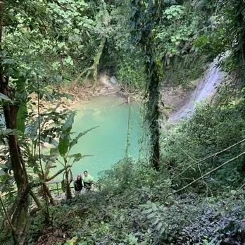 A lush green forest with a waterfall flowing into a turquoise pool, with two people standing on the edge of the water.