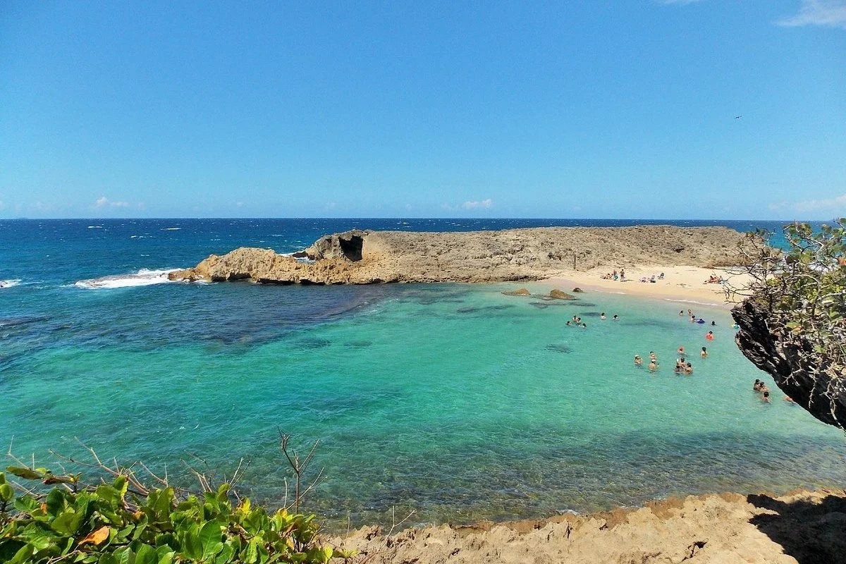 People swimming and relaxing in a small, shallow turquoise lagoon surrounded by rocky shores on a sunny day.
