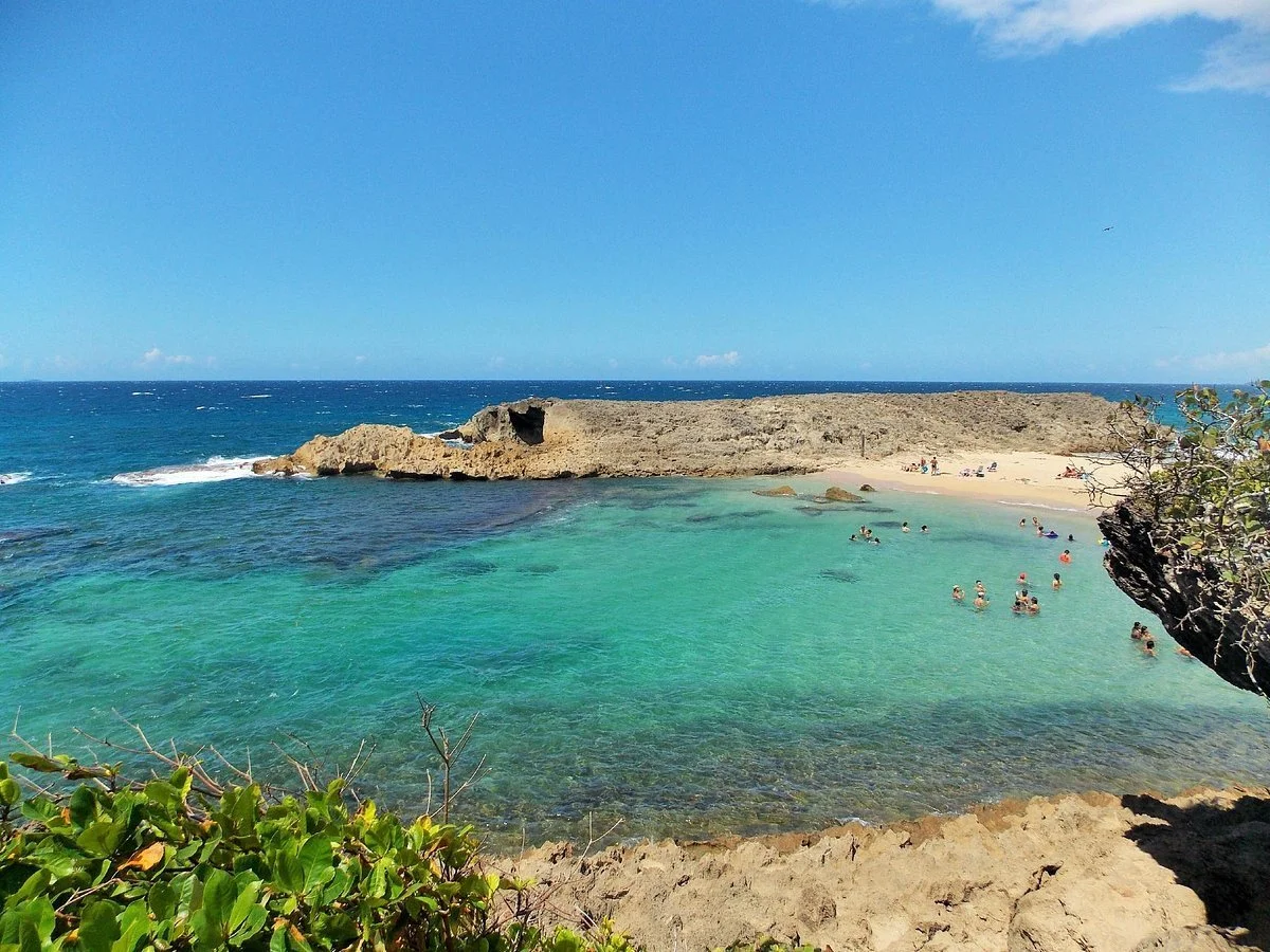 A coastal scene with a sandy beach, rocky outcrop, clear turquoise water, and people swimming and relaxing on the shore under a blue sky.