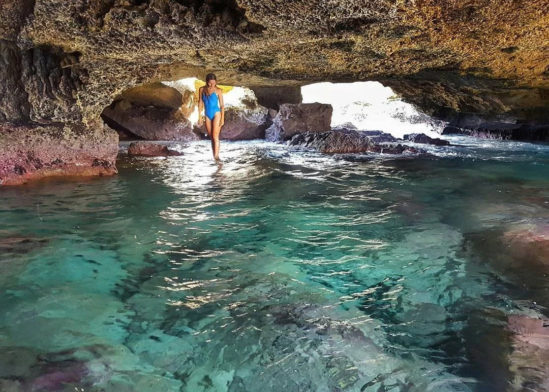 A woman in a blue swimsuit wades through clear turquoise water inside a rocky cave with an opening showing bright light outside.