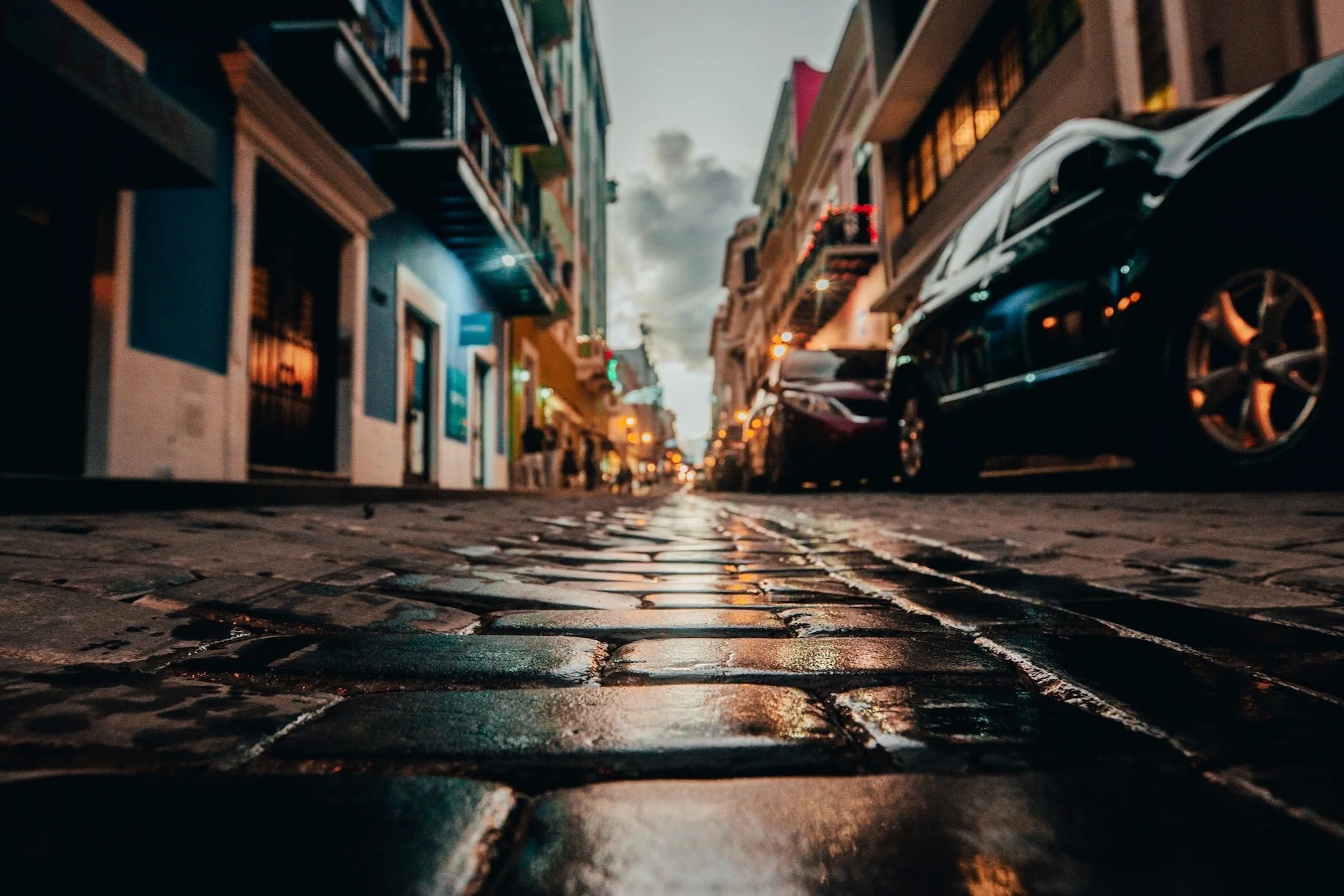Low-angle view of a city street with cobblestone pavement, parked cars, colorful buildings, and streetlights during dusk or evening.