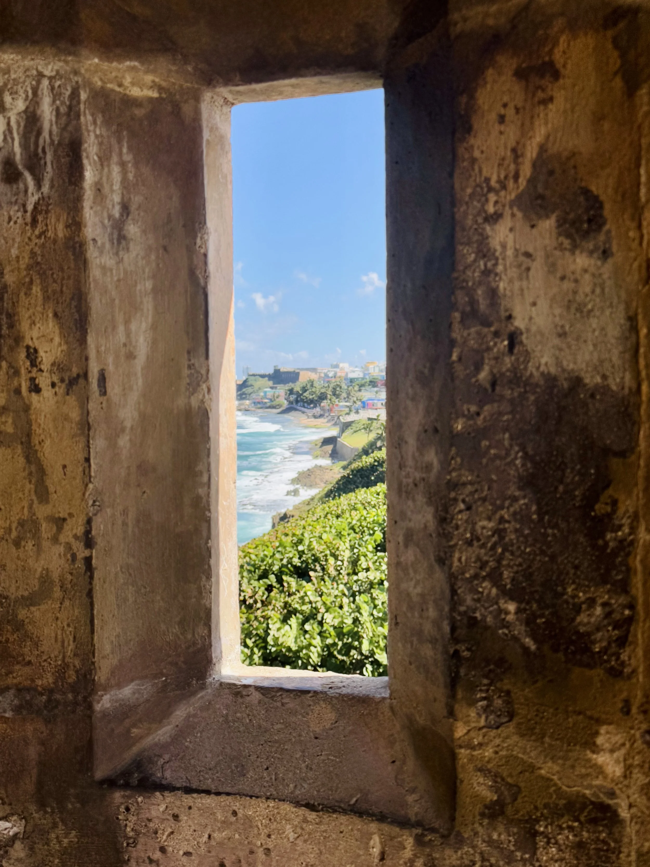 View of a coastal scene with blue ocean waves, greenery, and buildings seen through a narrow brick window.
