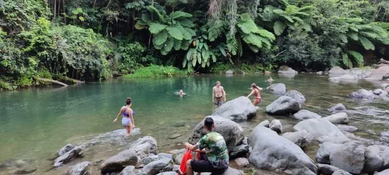 People swimming and relaxing in a clear river surrounded by lush green jungle with large rocks along the riverbank.