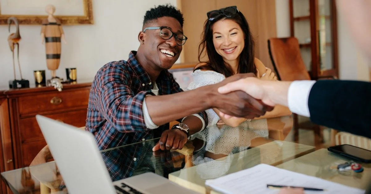 A smiling man sits across from a man in a suit at a desk, shaking his hand. A woman sits next to him, smiling.