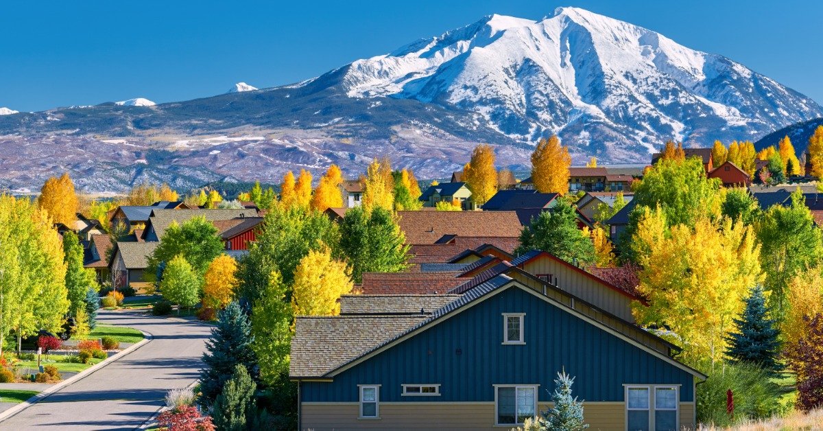 Trees with leaves radiating fall colors line a residential street with newer homes. A snow-capped mountain is in the background.