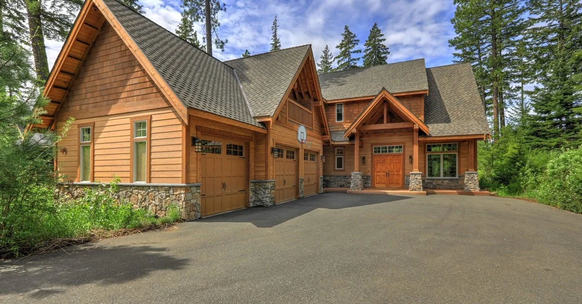 A large wooden house features pitched roofs, tall windows, a green lawn, and trees. It sits beneath a cloudy sky.