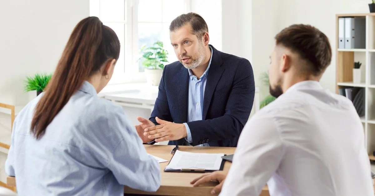 Two people listen as a real estate executive sits in his office, sternly explaining something. He sits behind a table.
