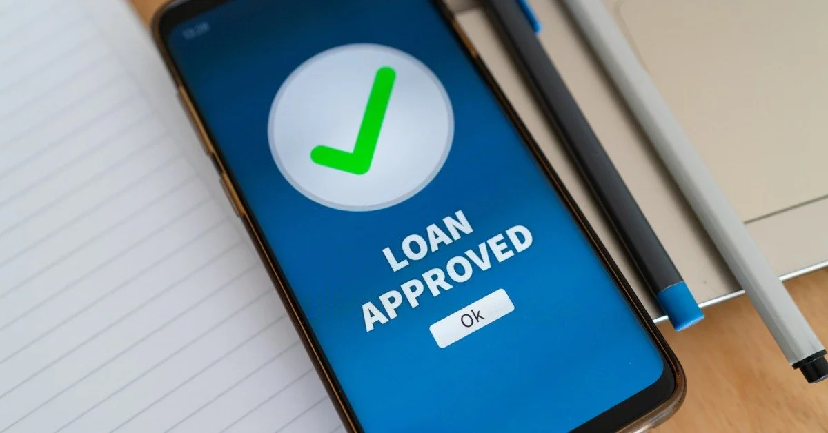 A smartphone sits next to two pens. Its screen reads the words "loan approved" with a giant checkmark in a circle.
