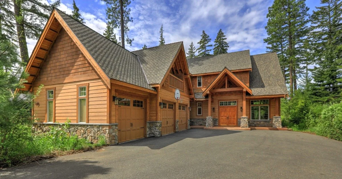 A home with cedar plank siding and stones sits on a property with oversized, mature evergreen trees and a driveway.