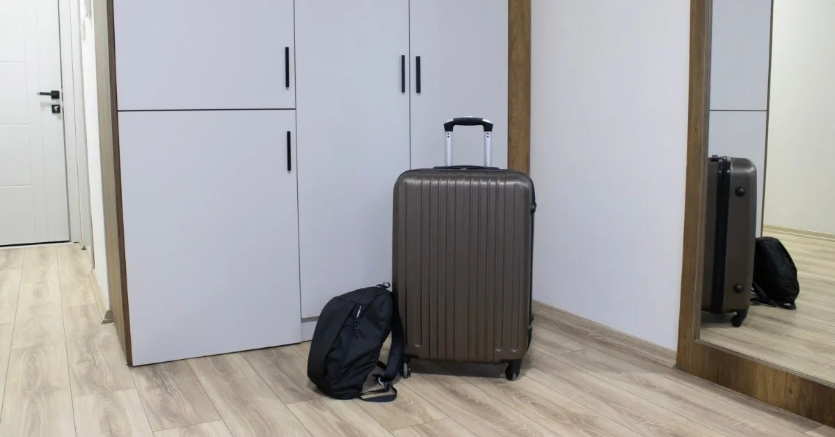 Black luggage with hard sides and on roller wheels has a black backpack leaning against it, sitting by the hallway storage.