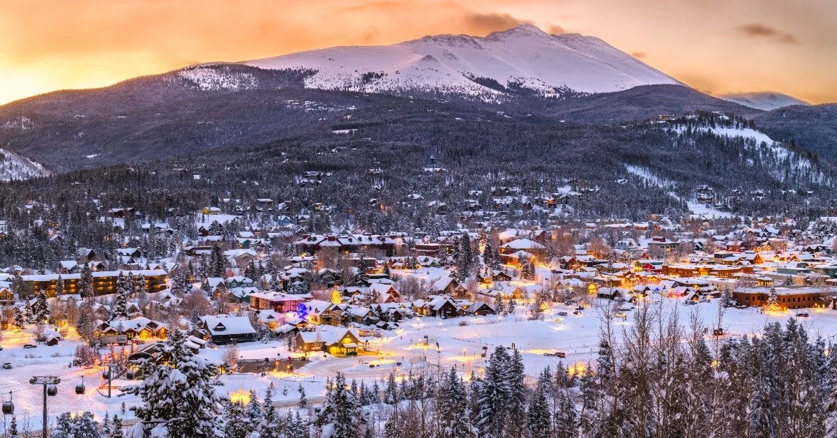 A beautiful town sits at the bottom of a mountain, covered in white snow. The town lights are on because it's dusk.