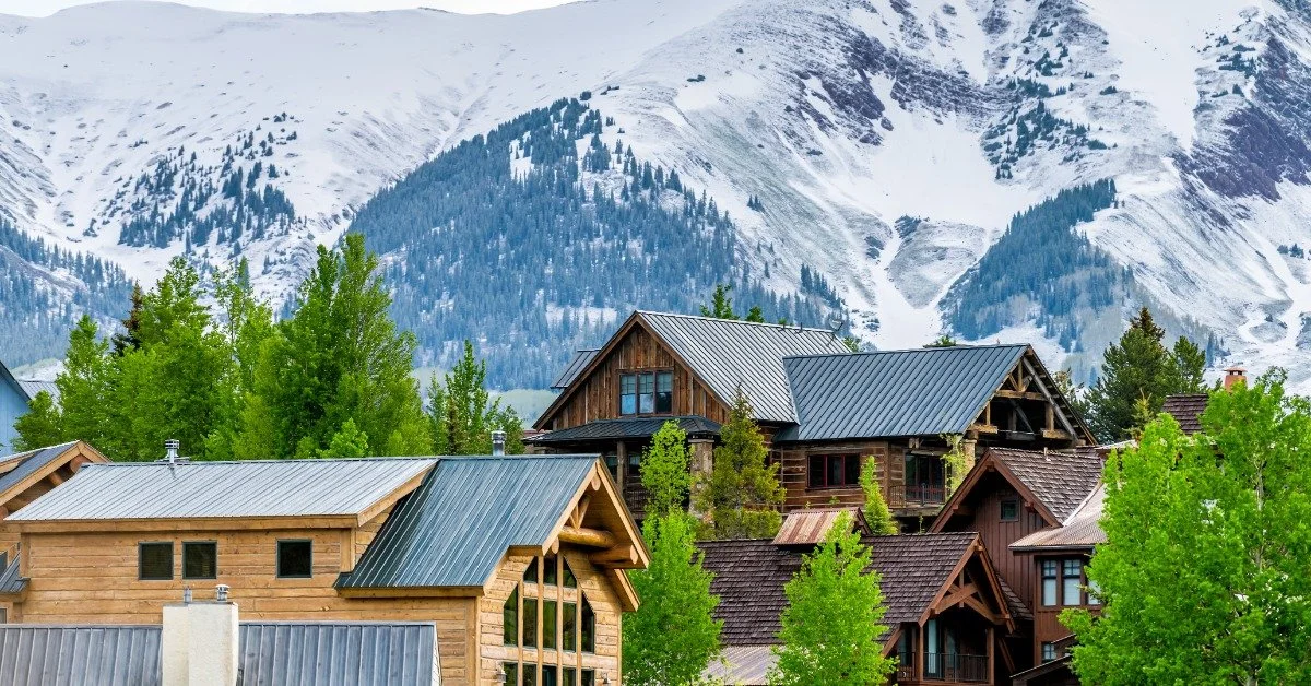 Beautiful, large homes are nestled among the trees with green leaves at the base of a snow-covered mountain.
