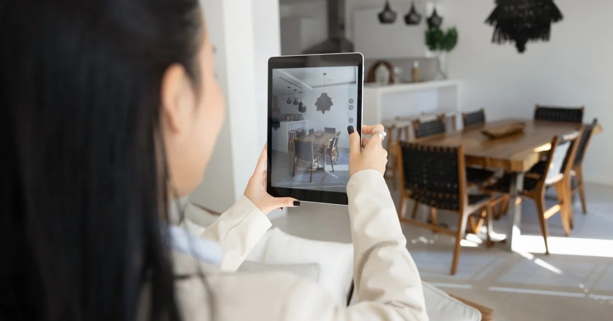 A woman takes a picture with a tablet of a dining room with a wooden table, chairs, and pendant lights.