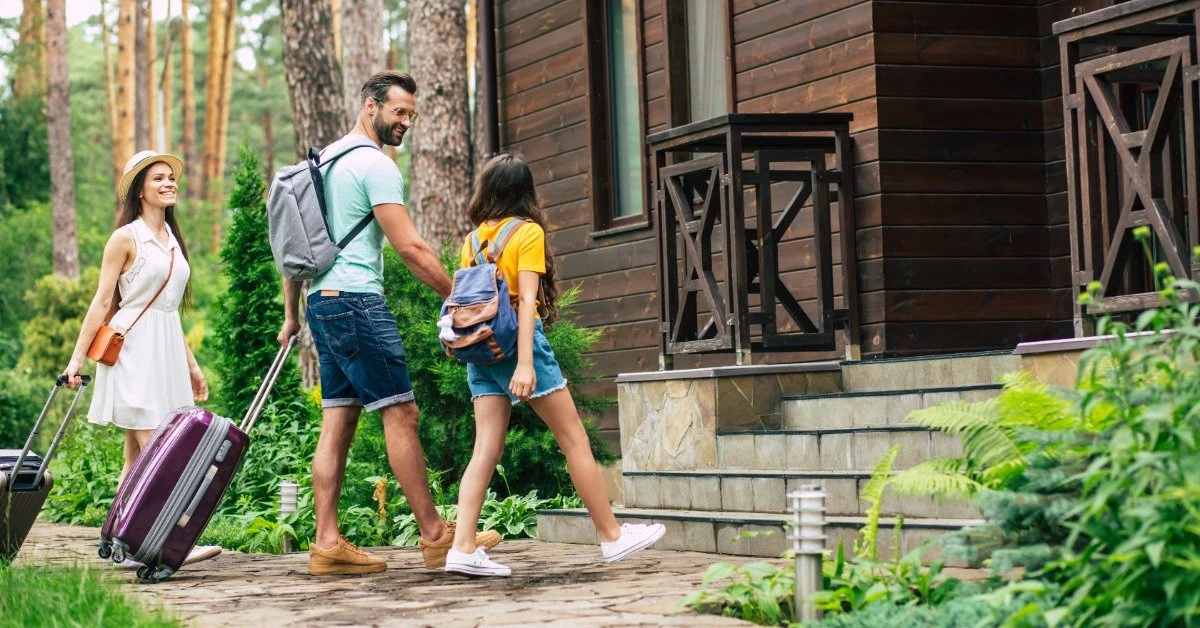 A father pulling luggage looks at his pre-teen daughter as they walk up the porch stairs. Mom is pulling luggage behind them.