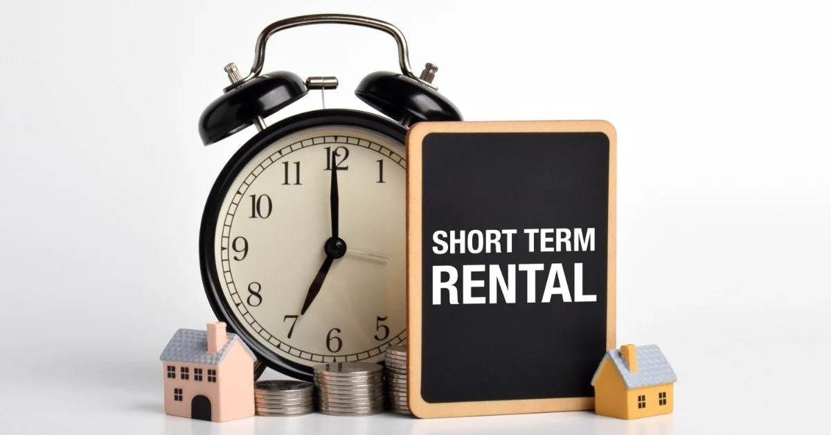 An alarm clock sits beside coin stacks, two small house models, and a sign reading "short term rental."