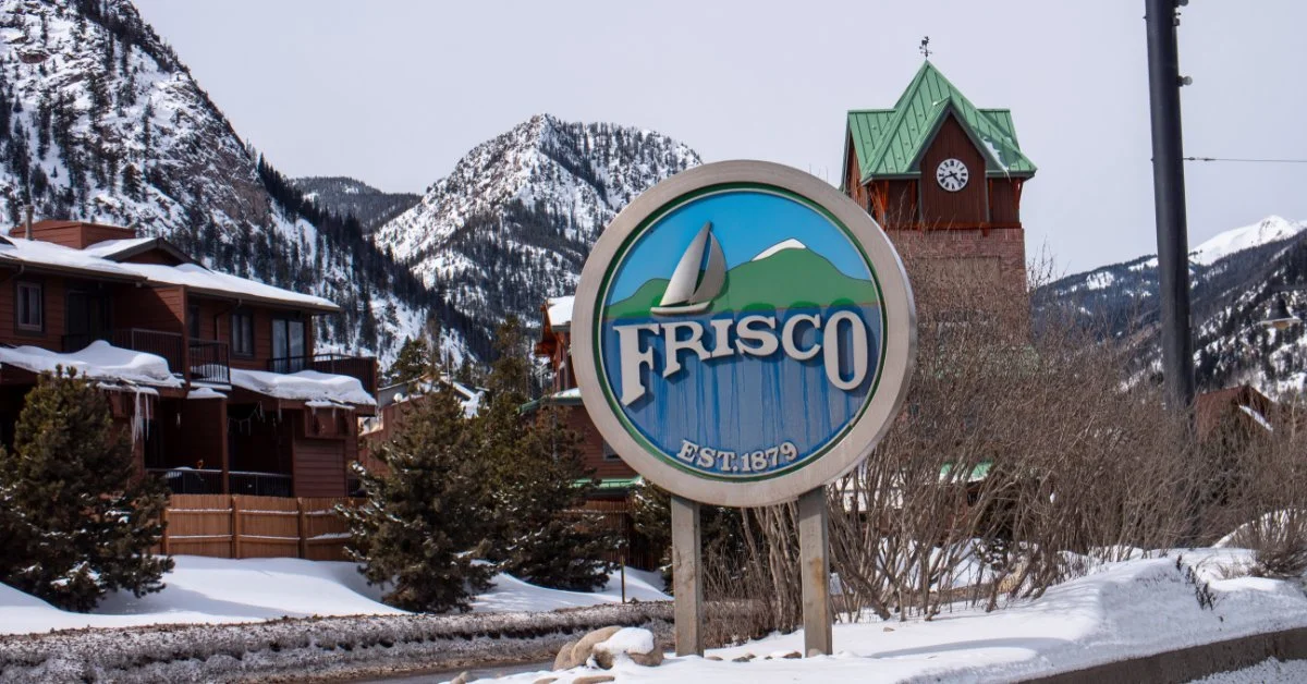 A Frisco, Colorado, welcome sign stands beside a snowy road with buildings and mountains in the background.