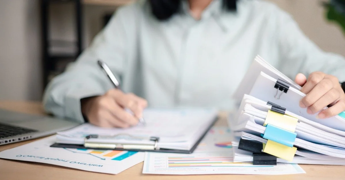A person sits at their desk, looking through a pile of documents bound together with clamps. They write on a clipboard.