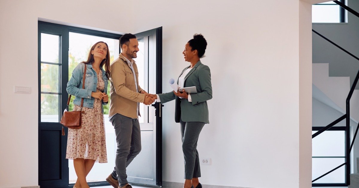A man and a woman are greeted by a female realtor at a home. The home has a modern black door and interior.
