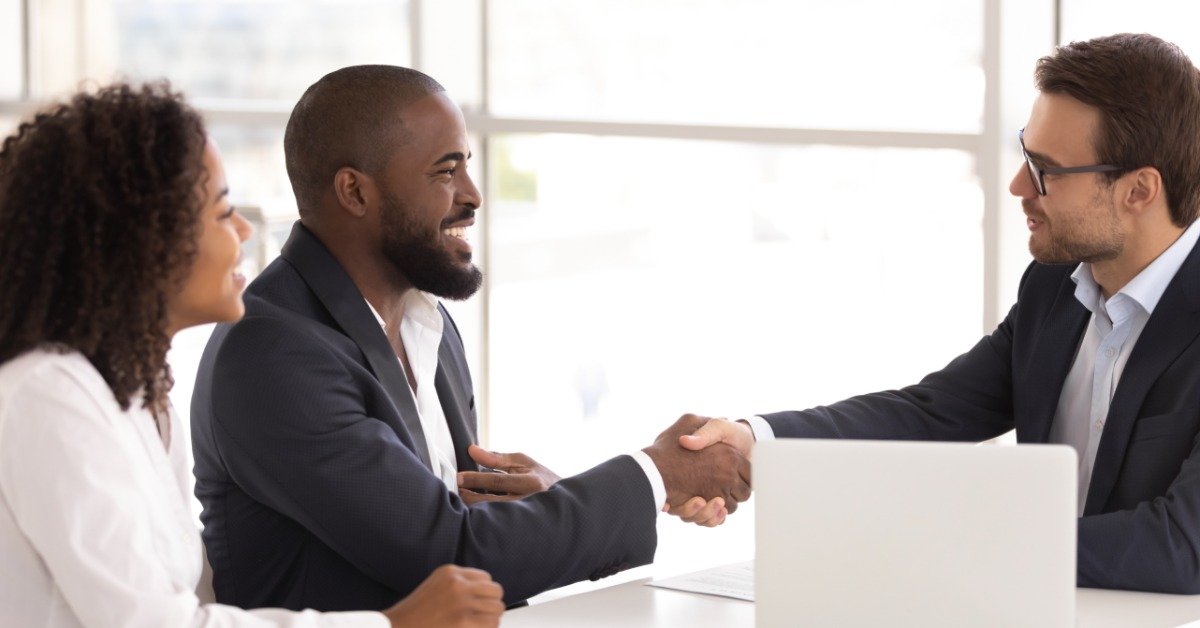 A happy couple sits across the desk from a man in his office. The husband shakes hands with the man.