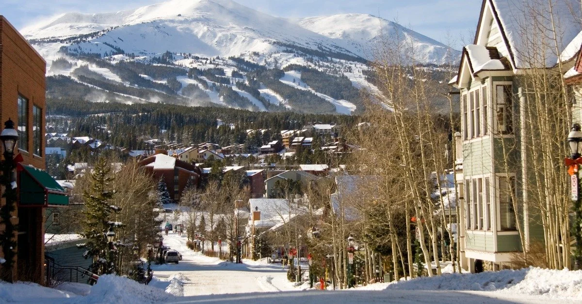 A snow-covered street is lined with homes, businesses, and trees. A beautiful mountain is in the background.