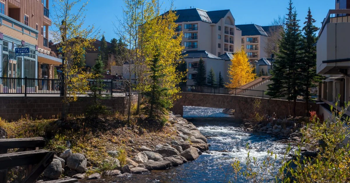 A rocky stream runs under a stone bridge between buildings and yellow autumn trees in Breckenridge, Colorado.