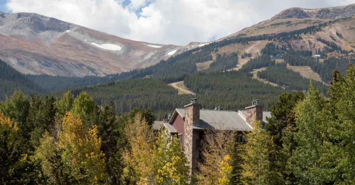 A home sits among several evergreen trees. A long range of tall mountains stands in the background behind it.