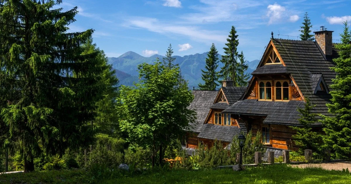 A large home with sharp roof lines sits on a property with a lot of mature trees and mountains in the background.