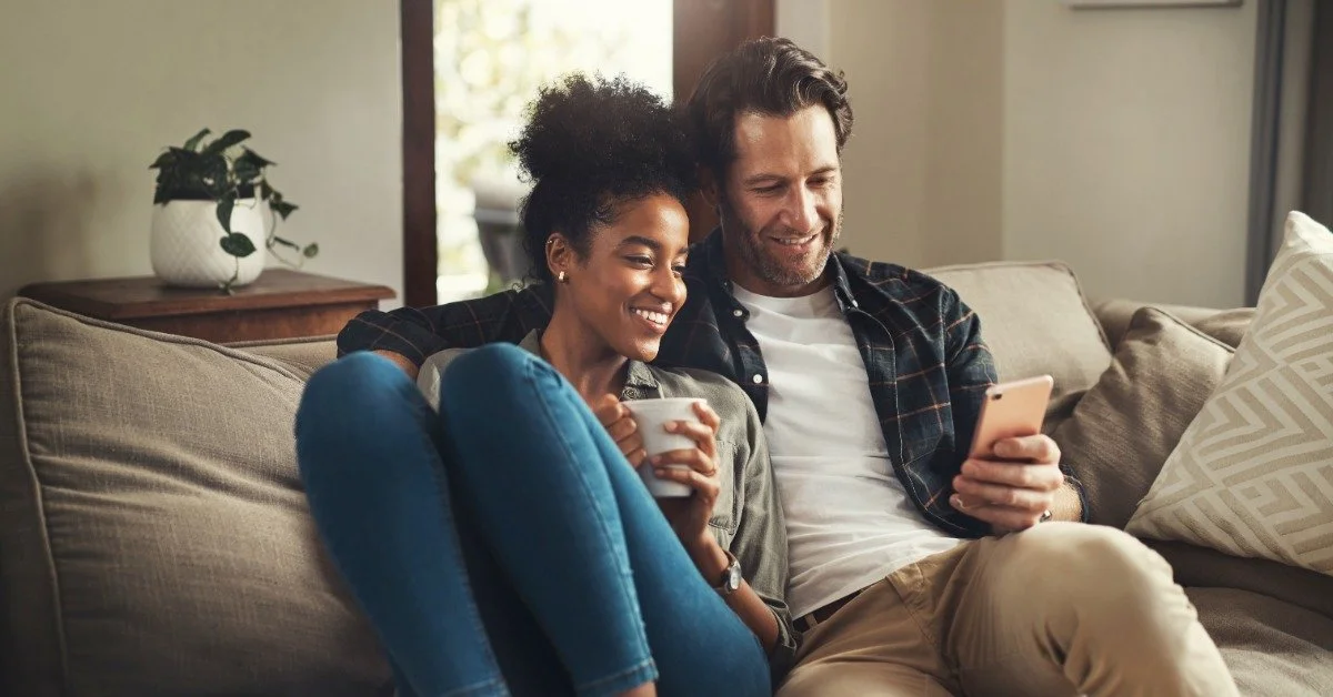 A man holds a phone while sitting on a couch. A smiling woman is snuggled into him, holding a mug and looking at the phone.