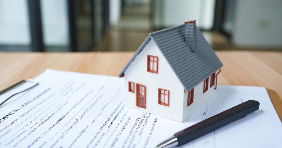 A small model of a house sits on a document on a clipboard that sits on a table in an office. A pen sits on the paper.