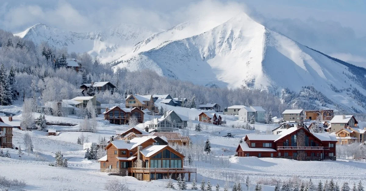 A snow-covered mountain range rises above a flat valley with roads, small houses, evergreen slopes, and a blue sky.