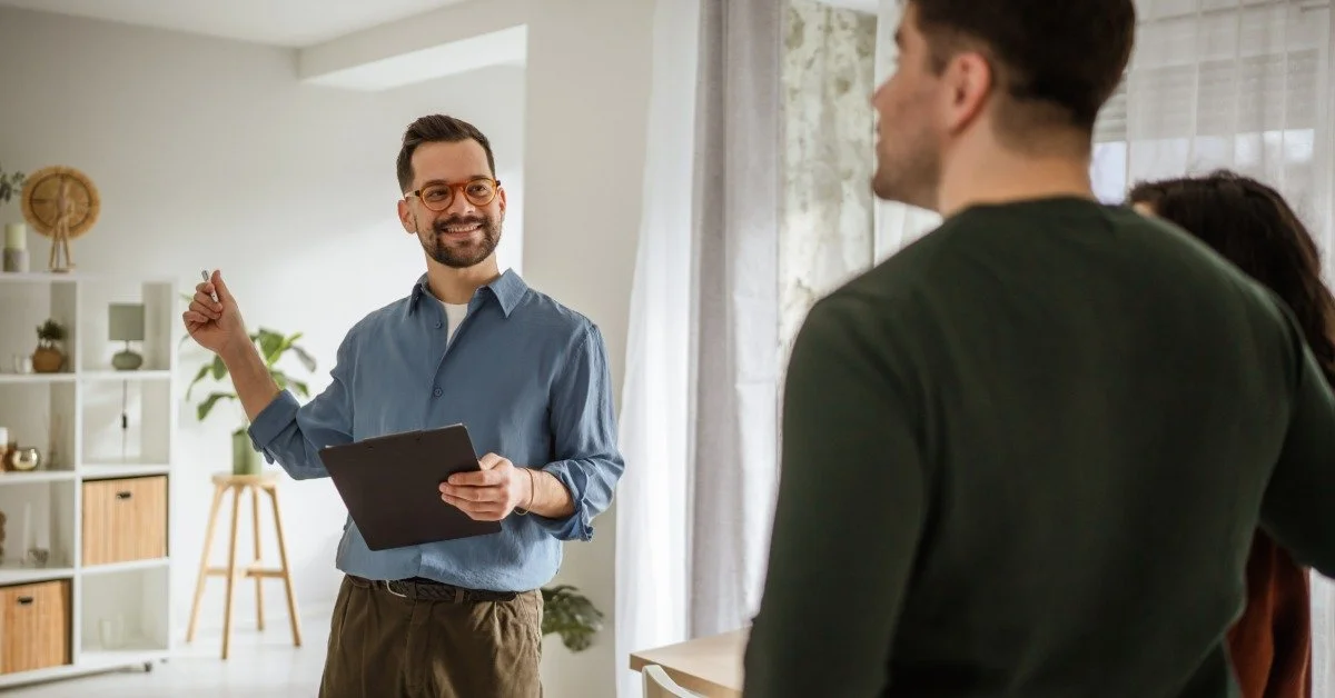 A real estate agent smiles as he holds a clipboard and shows a home to a couple. It features white walls.