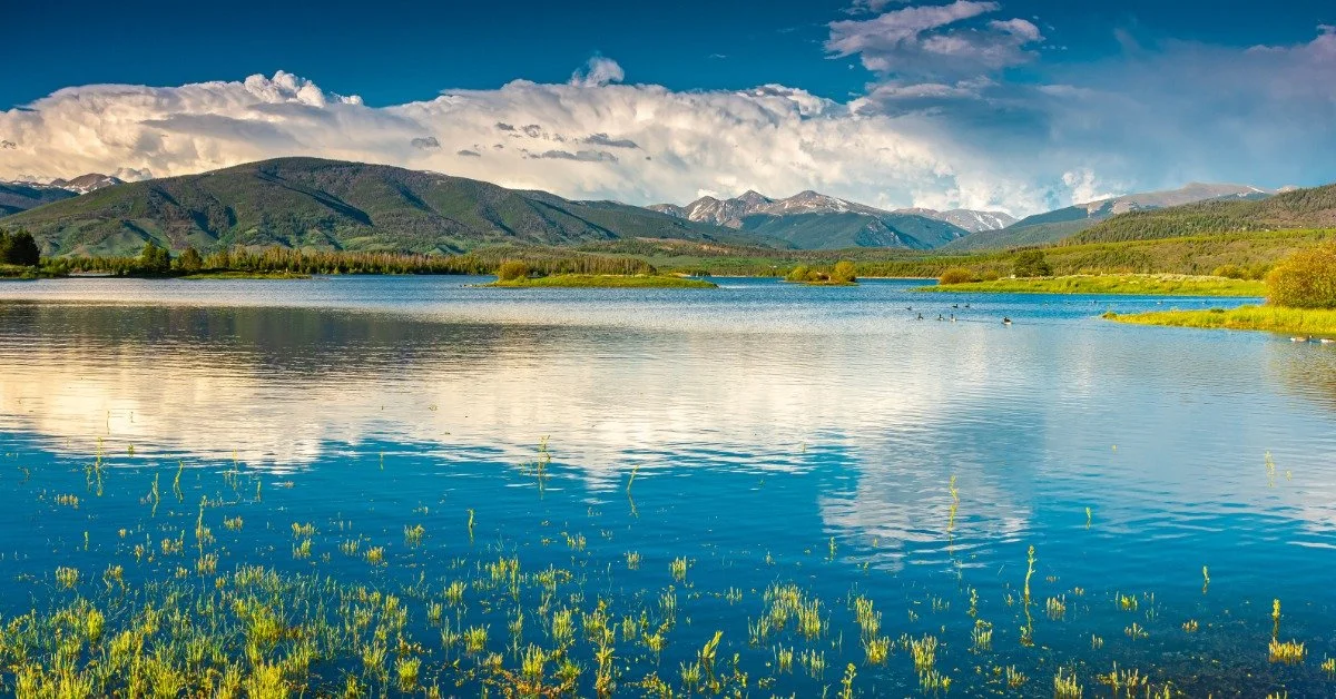 A large body of water sits at the bottom of large mountains. A blue sky with large, fluffy clouds is behind the mountains.