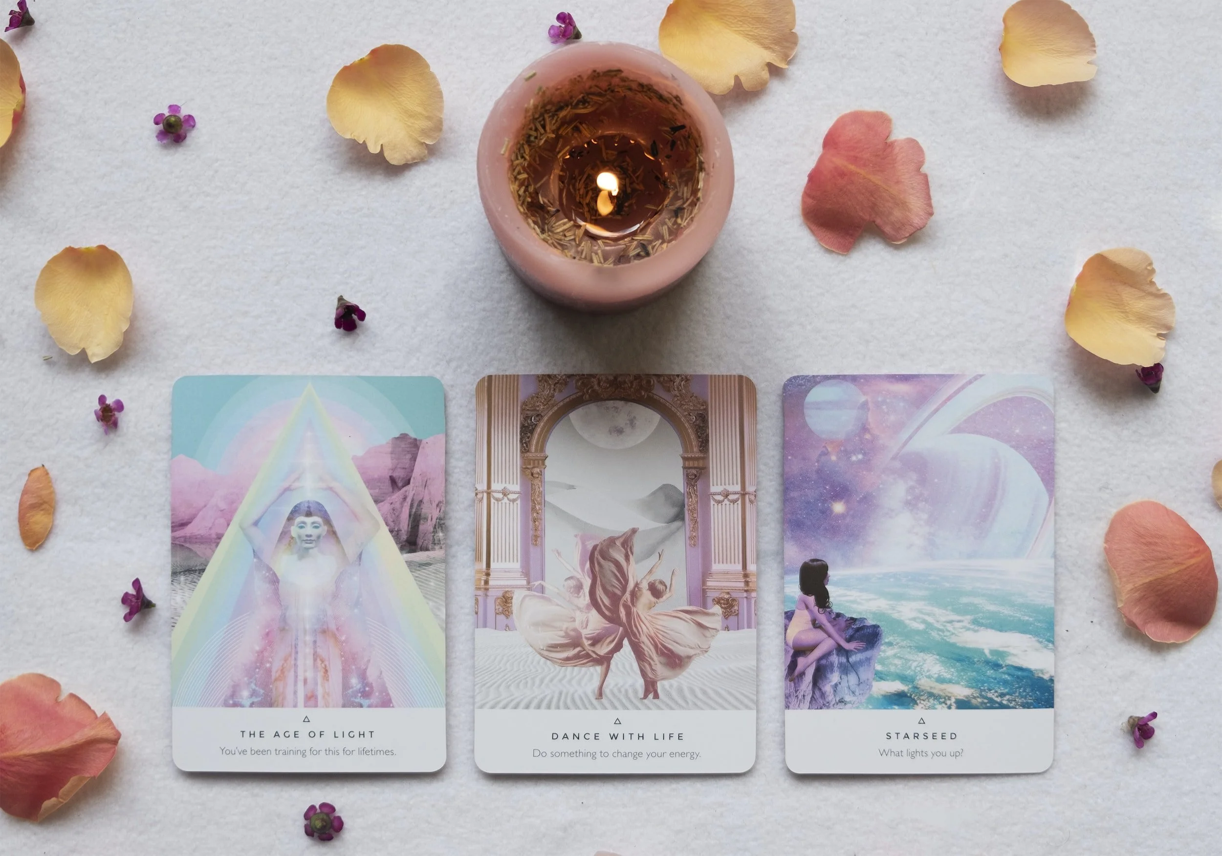 Three tarot or oracle cards laid out on a white surface with scattered flower petals and a glowing pink candle above them. The cards feature mystical and celestial themes, including a rainbow, a dancing figure, and a girl sitting on a rock looking at a cosmic sky.