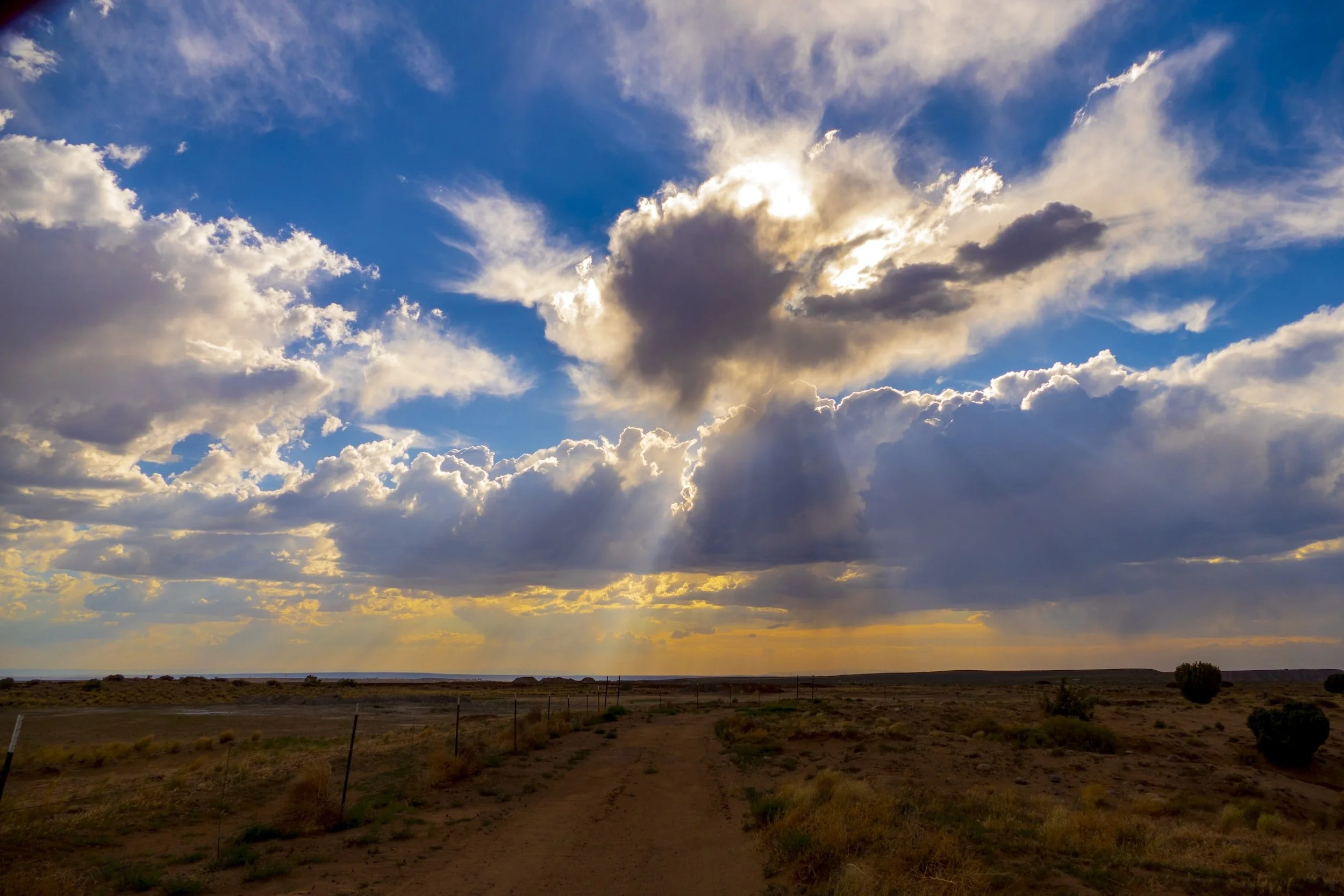 A dirt road leading into a vast open landscape under a dramatic sky filled with clouds during sunset.