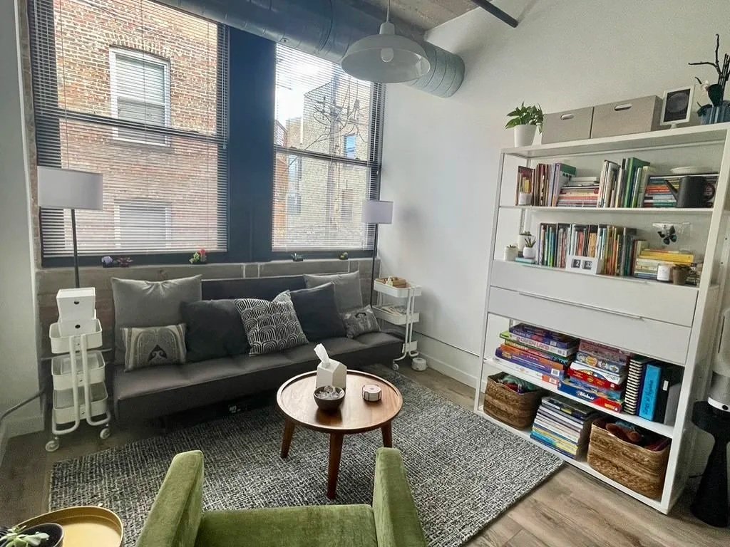 InnerBloom Therapy Practice cozy office space with modern couch, velvet green chair, throw pillows, shelving, and industrial style ceilings, with natural light and view of brick buildings in Chicago
