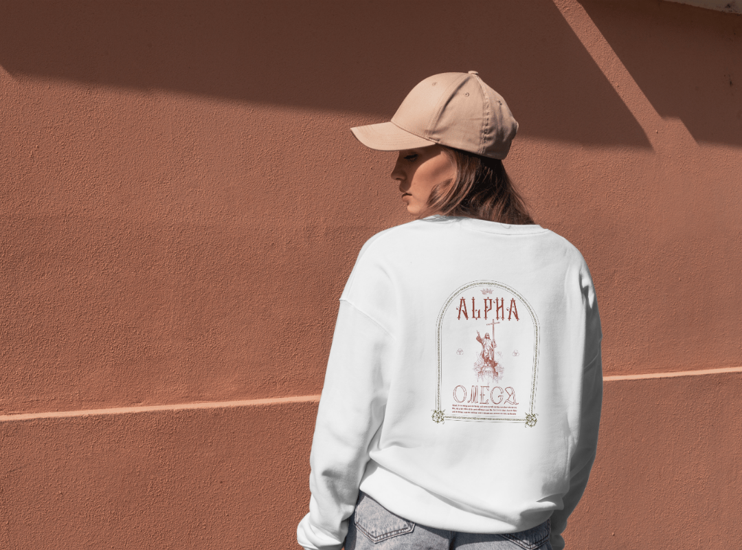 A woman wearing a beige baseball cap and a white sweatshirt with a design on the back, standing against a textured, reddish-brown wall with shadows.