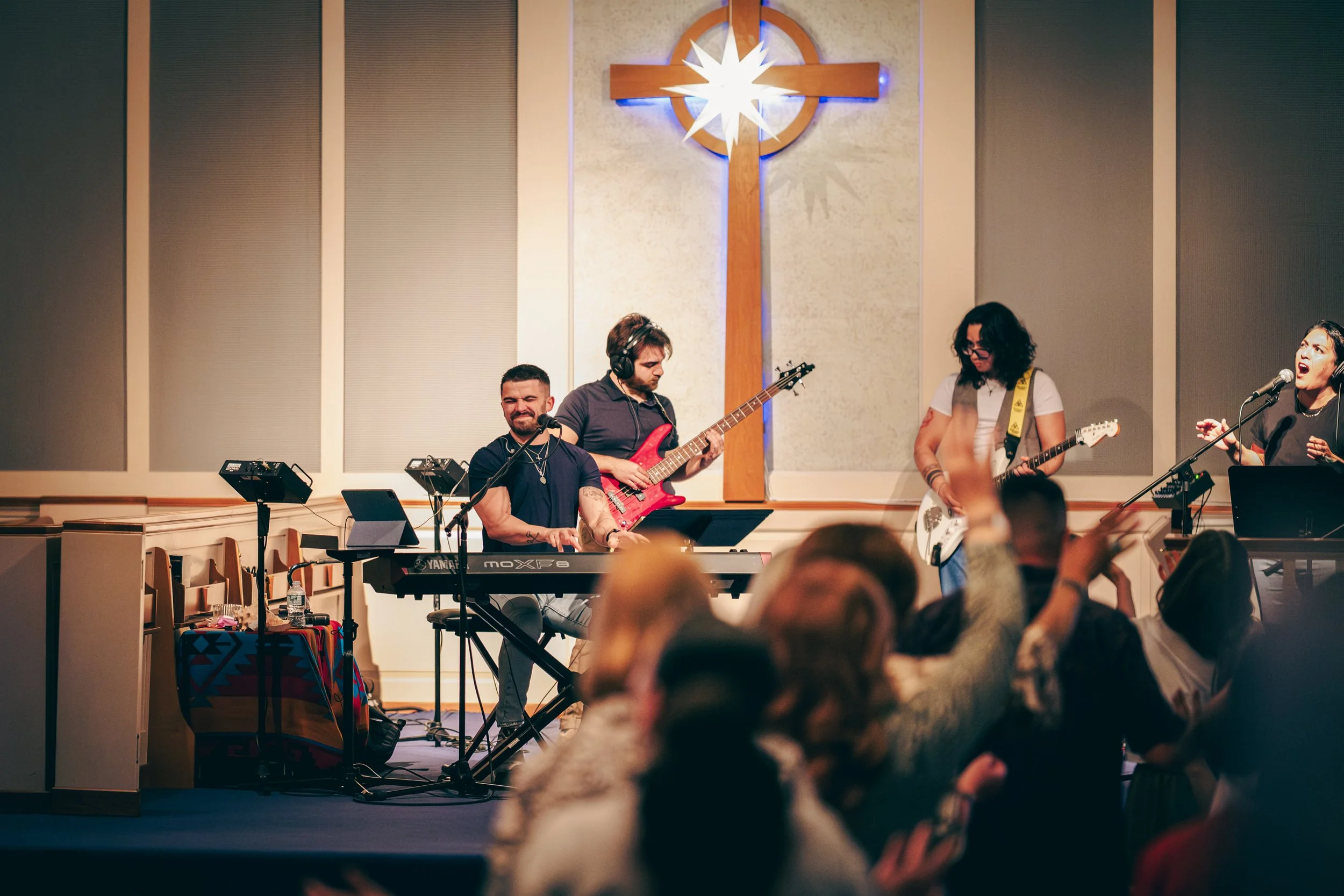 Stevie Mitchell and worship team performing on stage in a church setting with a cross and a star-shaped light behind them. The band includes a keyboardist, a guitarist, a bassist, and a singer.