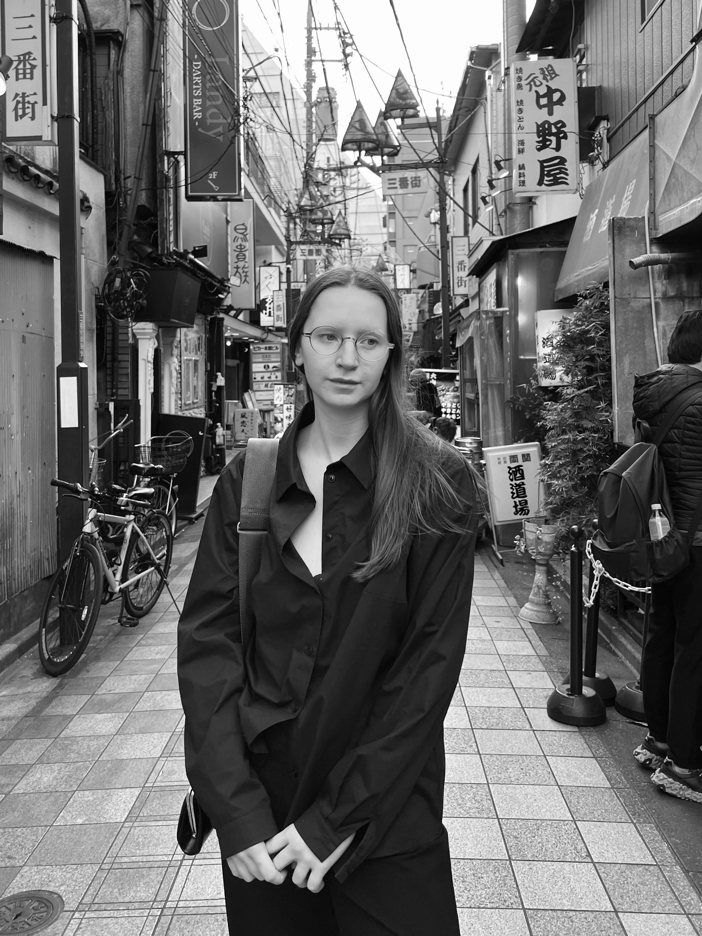 A woman with long hair and glasses wearing a black shirt standing on a busy street in Japan with signs and bicycles in the background.