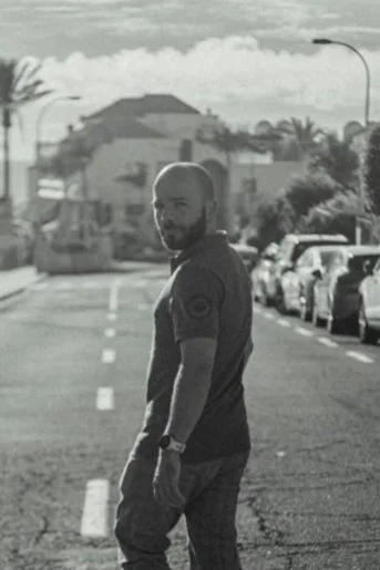 Man with a beard standing on a street in a neighborhood with parked cars, palm trees, and houses in the background, in black and white.