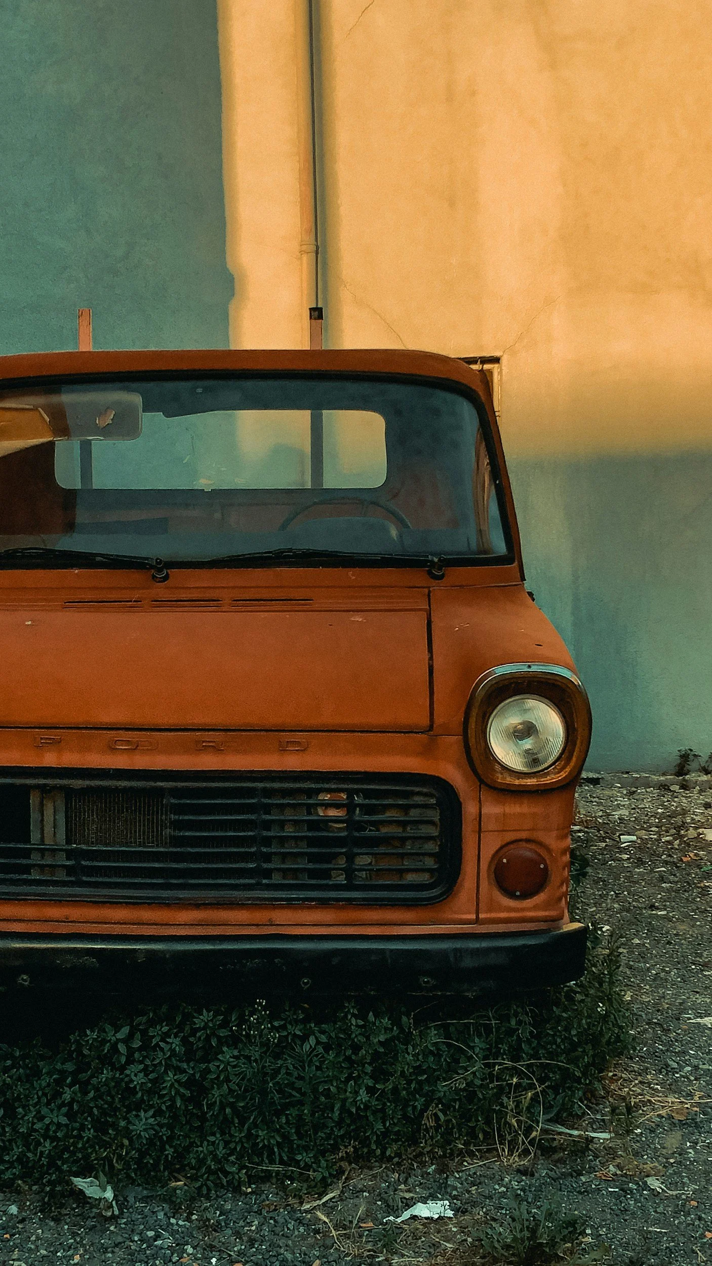 An old orange Ford vehicle parked with a cracked wall and overgrown plants in the foreground.