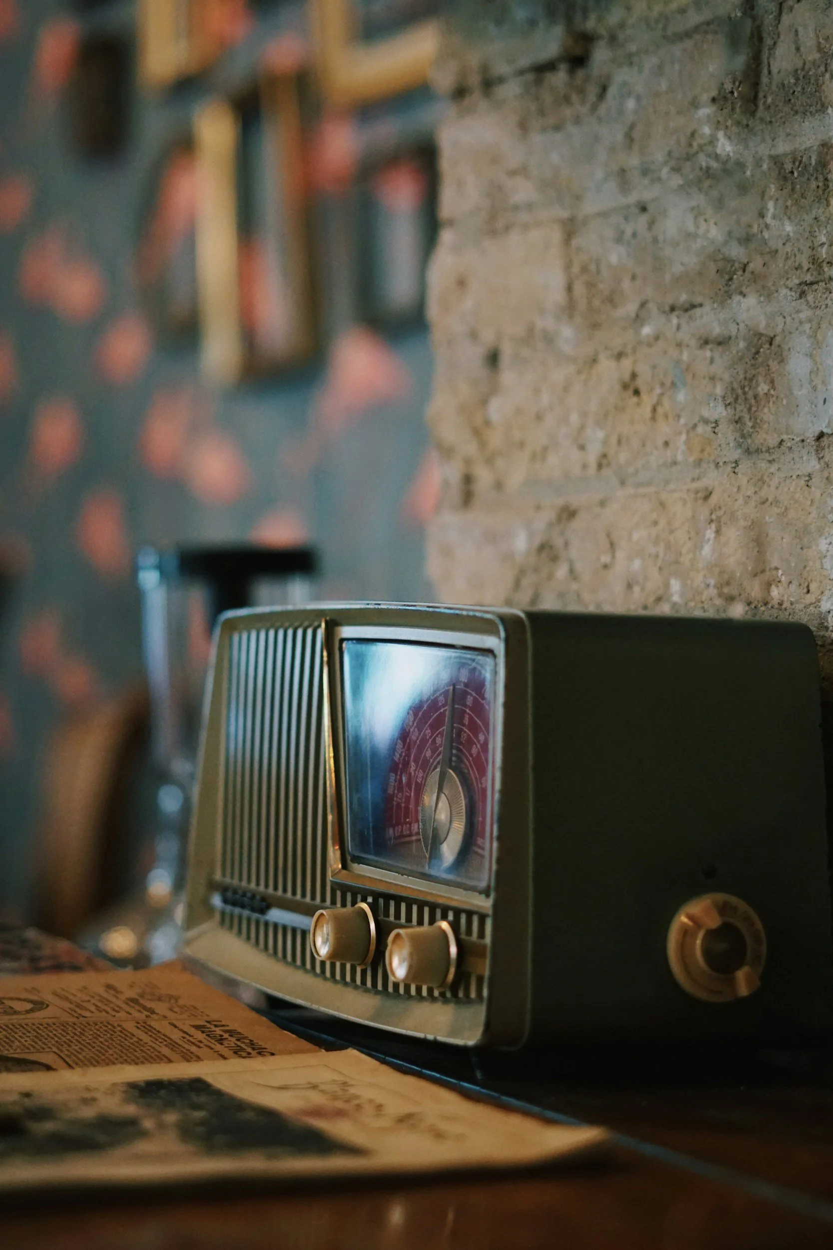 A vintage clock radio with two knobs sits on a table or counter, with a newspaper and a brick wall in the background.