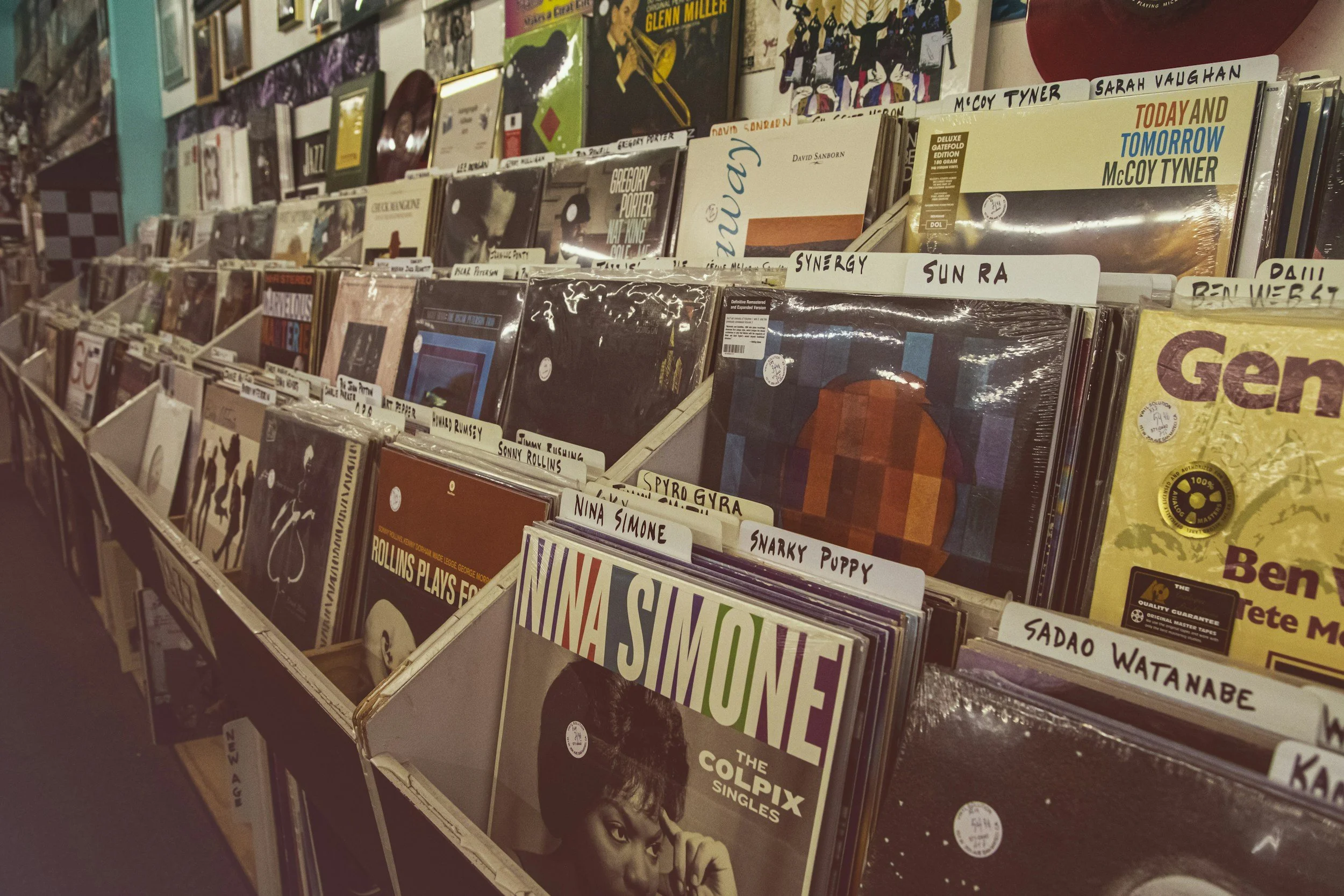 A display of vinyl records in a store, organized in wooden bins with handwritten labels. The covers feature various artists and album titles, with some recognizable names like McCoy Tyner and The Colpix Singles.