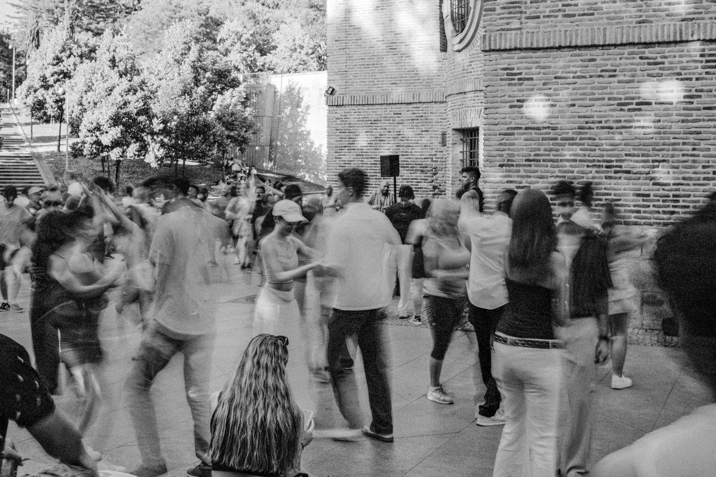People dancing and socializing indoors near a brick wall and large windows, with some sitting and others standing or dancing.