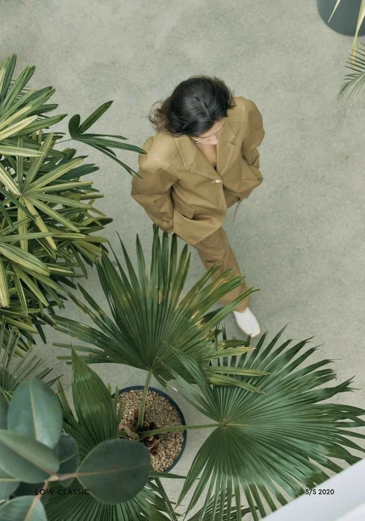 A woman in a tan suit and white shoes standing on a gray carpet floor, viewed from above near green potted plants.